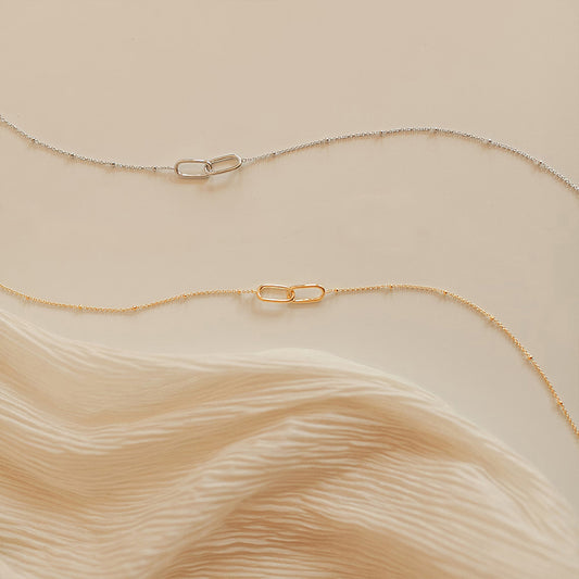 Pair of gold and silver infinity link pendant necklaces with delicate stainless steel chains on soft beige fabric.