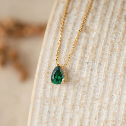 Gold chain necklace with green teardrop birthstone crystal pendant, displayed on textured background.