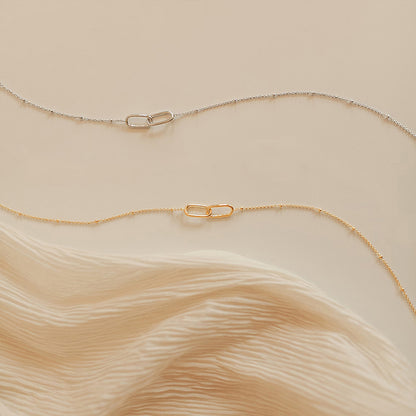 Pair of gold and silver infinity link pendant necklaces with delicate stainless steel chains on soft beige fabric.